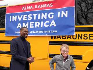 El administrador Regan y un estudiante pequeño sonrientes y de pie frente a un autobús escolar